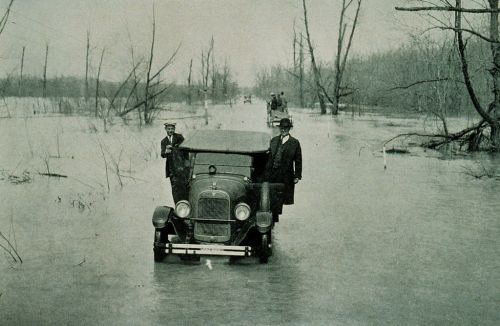 Two men stand beside a vintage car in a flooded area, with bare trees in the background and water covering the ground.