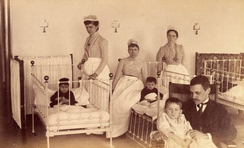 Historical photo of caregivers and children in a nursery, featuring cribs and a family setting from the late 19th century.