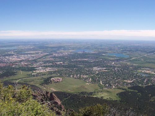 A panoramic view of a valley with a city, lakes, and green hills under a clear blue sky.