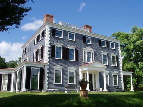 A large, historic house with blue siding, white trim, and a front porch, surrounded by green trees and a clear blue sky.