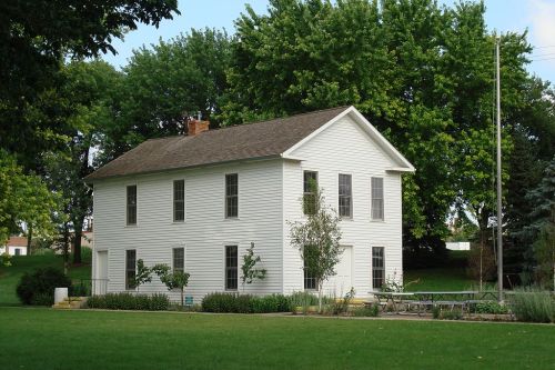 A white two-story house surrounded by green trees and a grassy area, with a small garden in front.