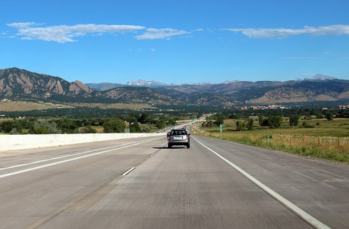 A highway stretches into the distance, flanked by mountains and clear blue skies. A car drives ahead on the road.