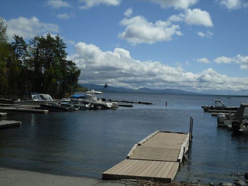 A serene lake scene with boats docked, a wooden pier, and clouds reflecting in the water under a blue sky.