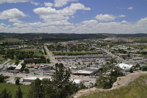 Aerial view of a small town with buildings, roads, and green spaces under a partly cloudy sky.