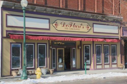 A charming storefront of DeFluri's Fine Chocolates, featuring large windows and a red awning, with snow on the ground.