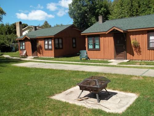 Two wooden cabins with green roofs beside a fire pit on a grassy area, under a blue sky with scattered clouds.