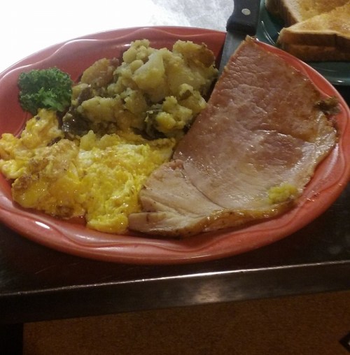 A red plate with scrambled eggs, ham, mashed potatoes, and a piece of broccoli.