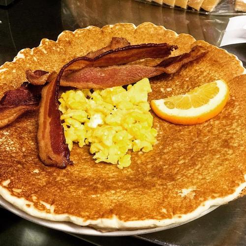 A large pancake topped with scrambled eggs, crispy bacon, and a slice of orange.