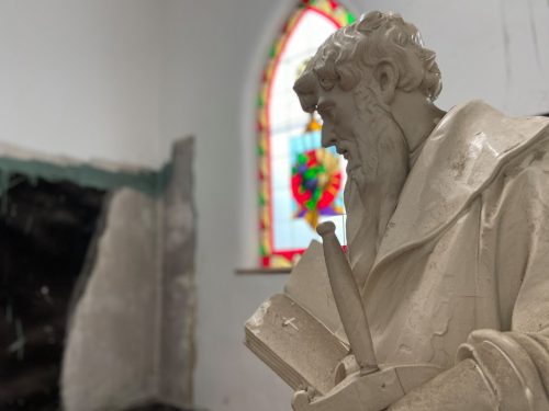 A close-up of a white statue of a bearded man holding a book, with a stained glass window in the background.
