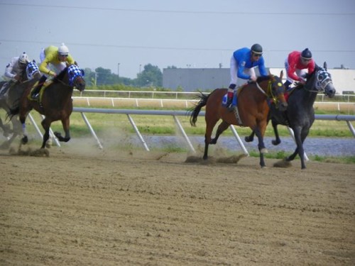Four horses racing on a dirt track, with jockeys in colorful uniforms, dust flying as they compete.