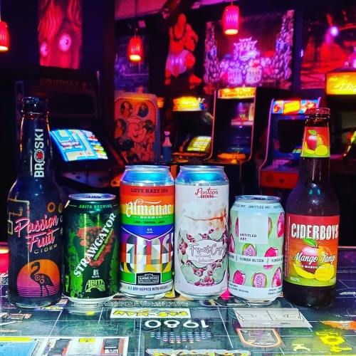 A colorful display of craft beers and ciders on a table, with arcade games in the background.