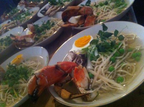 A close-up of a bowl of noodle soup with crab claws, peas, and herbs, surrounded by additional bowls of the same dish.