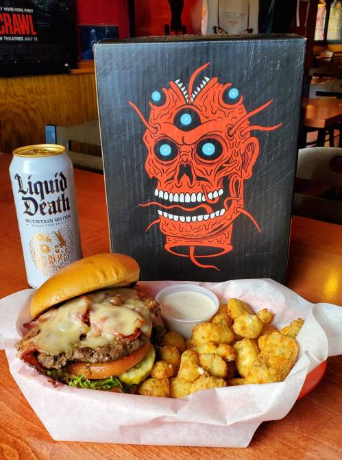 A burger with melted cheese and toppings, served with crispy bites and a can of Liquid Death, next to a colorful skull menu.