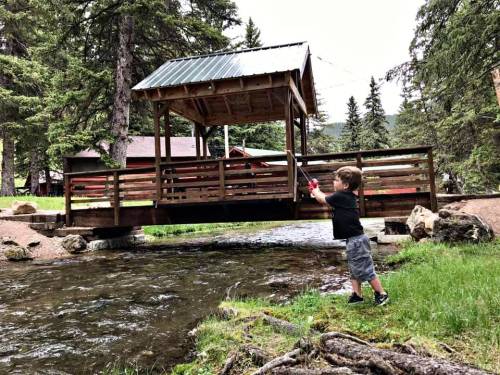 A young child stands by a stream, aiming a toy water gun at a wooden bridge surrounded by trees.