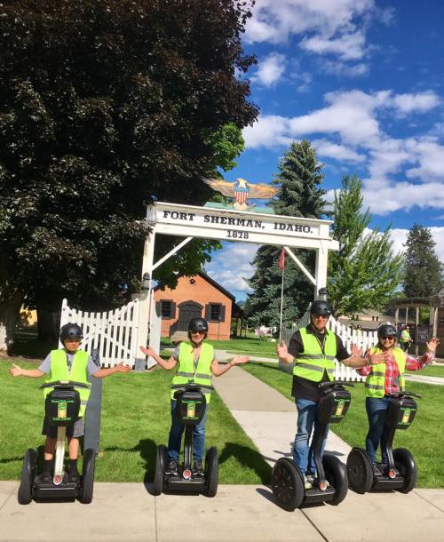 Four people in safety vests on Segways pose in front of a historic sign at Fort Sherman, Idaho, with trees and blue sky.