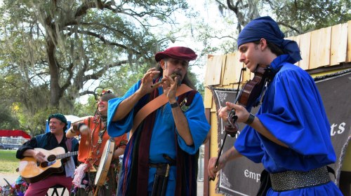 A group of musicians in colorful costumes perform outdoors, playing guitar, flute, and violin under trees.