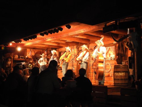 A country band performs on stage in a rustic venue, with an audience enjoying the music. Warm lighting enhances the atmosphere.