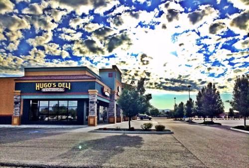 A deli building with a sign reading "Hugo's Deli" under a cloudy sky, surrounded by trees and an empty parking lot.