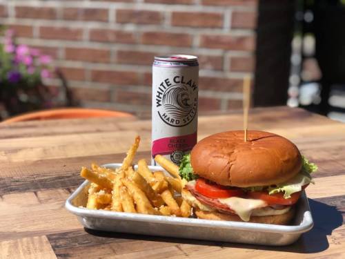 A burger with lettuce, tomato, and cheese, served with fries and a can of Black Cherry White Claw on a wooden table.