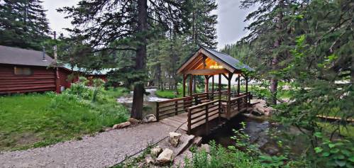 A wooden bridge leads to a cozy cabin surrounded by tall trees and a flowing stream in a serene forest setting.
