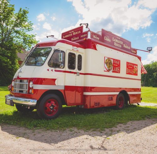 A vintage food truck painted red and white, serving lobster rolls, parked on a grassy area with trees in the background.