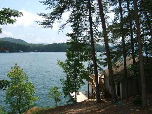 A serene lake view surrounded by trees, with a wooden dock extending into the water and a house nearby.