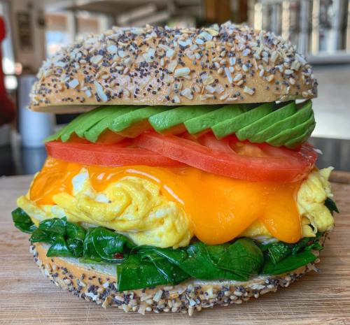A bagel sandwich with poppy seeds, filled with spinach, scrambled eggs, cheddar cheese, tomato, and avocado slices.