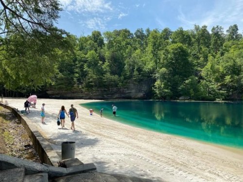 A sandy beach by a clear blue lake, surrounded by lush green trees, with people enjoying the outdoors.