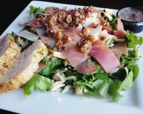 A fresh salad with greens, orzo, prosciutto, nuts, cheese, and a side of dressing, served with a slice of bread.