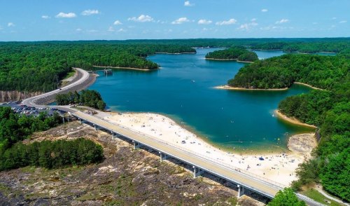 Aerial view of a lake surrounded by green forests, with a sandy beach and a road running alongside it.