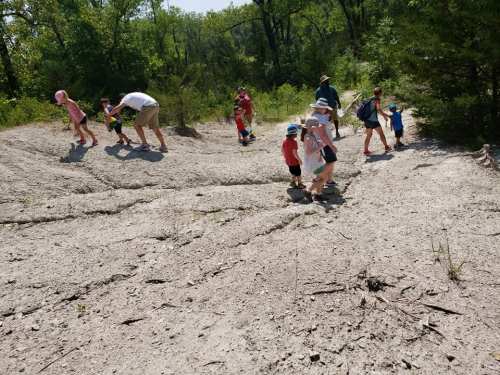 A group of children and adults exploring a dry, sandy area with trees in the background on a sunny day.