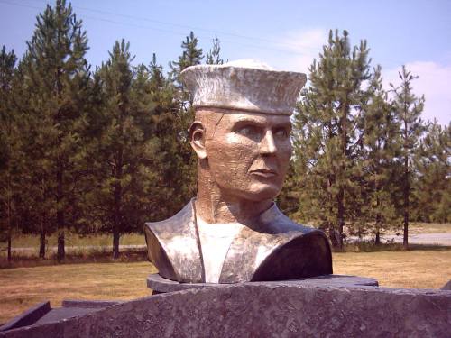 A bronze bust of a sailor in a naval uniform, set against a backdrop of trees and blue sky.