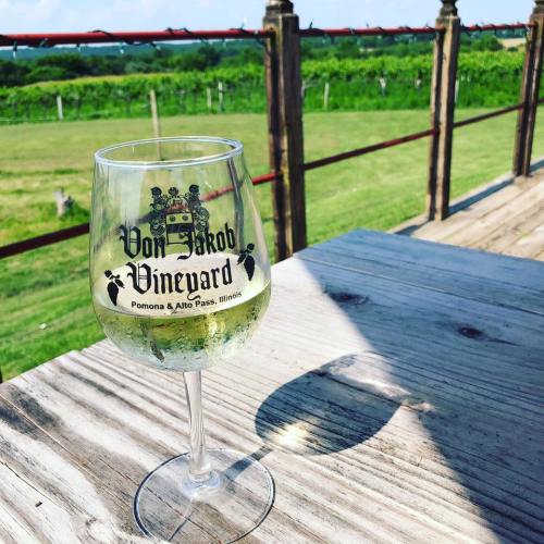 A glass of white wine from Von Jakob Vineyard sits on a wooden table, with vineyards in the background under a blue sky.