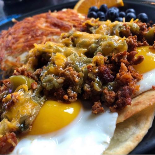 A close-up of a hearty breakfast plate featuring eggs, chorizo, cheese, hash browns, and fresh fruit.