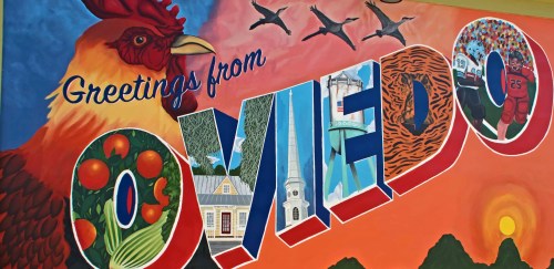 Colorful mural reading "Greetings from Oviedo," featuring a rooster, nature, and local landmarks.