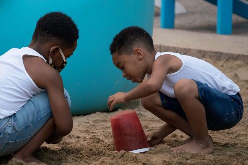 Two young boys play in a sandbox, focused on building with a red bucket. One wears a mask, both are in casual summer clothes.
