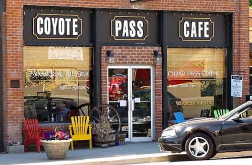 Exterior of Coyote Pass Cafe with colorful chairs, flower pots, and an open sign on a brick building.