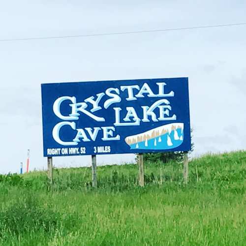 Sign for Crystal Lake Cave, located 3 miles right on Highway 52, with a blue background and decorative lettering.
