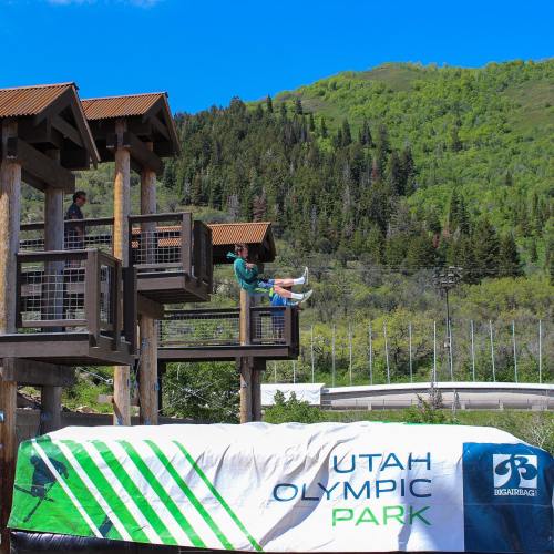 A person prepares to jump from a platform at Utah Olympic Park, surrounded by green mountains and blue skies.