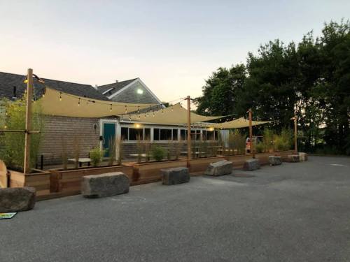 Outdoor seating area with wooden deck, shade sails, and string lights, surrounded by greenery and a building in the background.