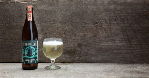 A bottle of Stormalong Legendary Dry beer next to a glass filled with the beverage, set against a wooden background.