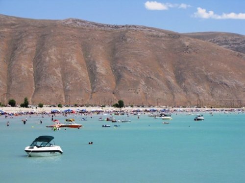 A sunny beach scene with people, boats, and a backdrop of brown hills under a clear blue sky.