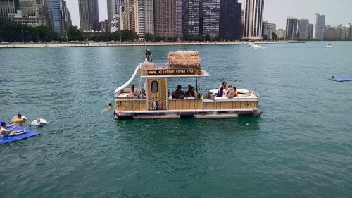 A party boat with a thatched roof floats on a city lake, surrounded by swimmers and skyscrapers in the background.
