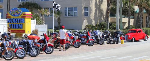 A row of motorcycles parked by a beachside road, with a vintage red car and a sign for "North Turn" nearby.
