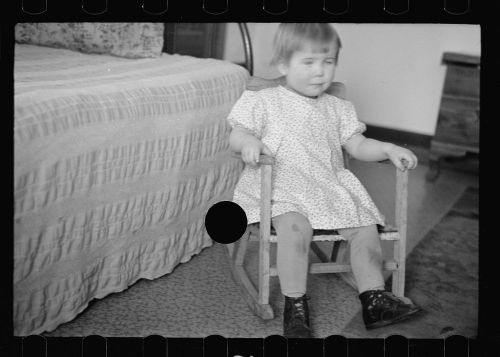 A young child in a floral dress sits on a small rocking chair, with a blurred expression, in a cozy room.
