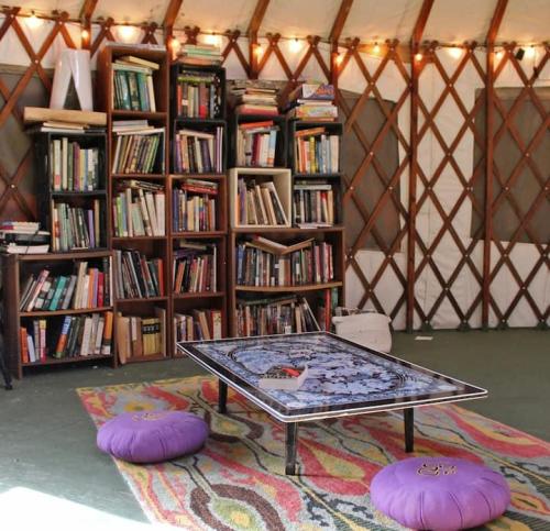 Cozy yurt interior with a bookshelf, colorful rug, low table, and purple cushions, illuminated by string lights.