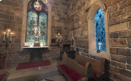 A cozy chapel with stone walls, stained glass windows, candles, and wooden pews.