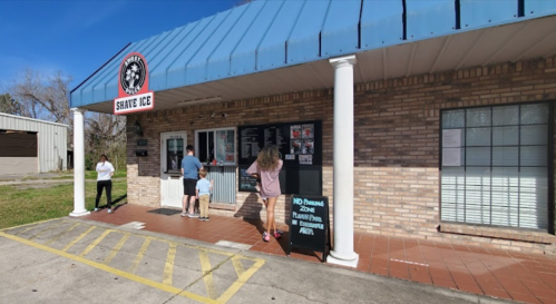 A brick building with a "Shave Ice" sign; people are ordering at the window on a sunny day.