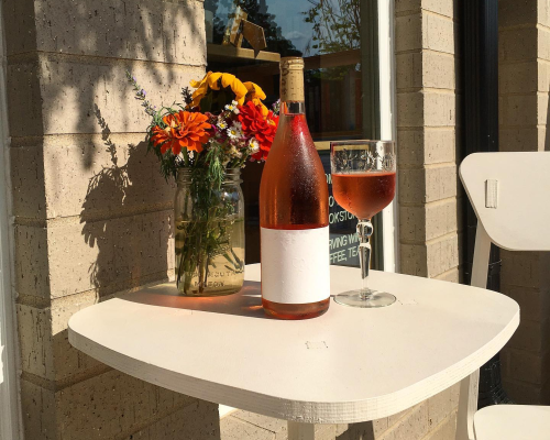 A bottle of rosé wine and a glass sit on a white table beside a vase of colorful flowers.