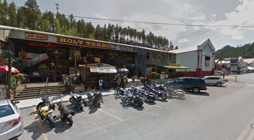 A rustic building with "Holy Terror" sign, surrounded by trees, featuring outdoor seating and parked motorcycles.